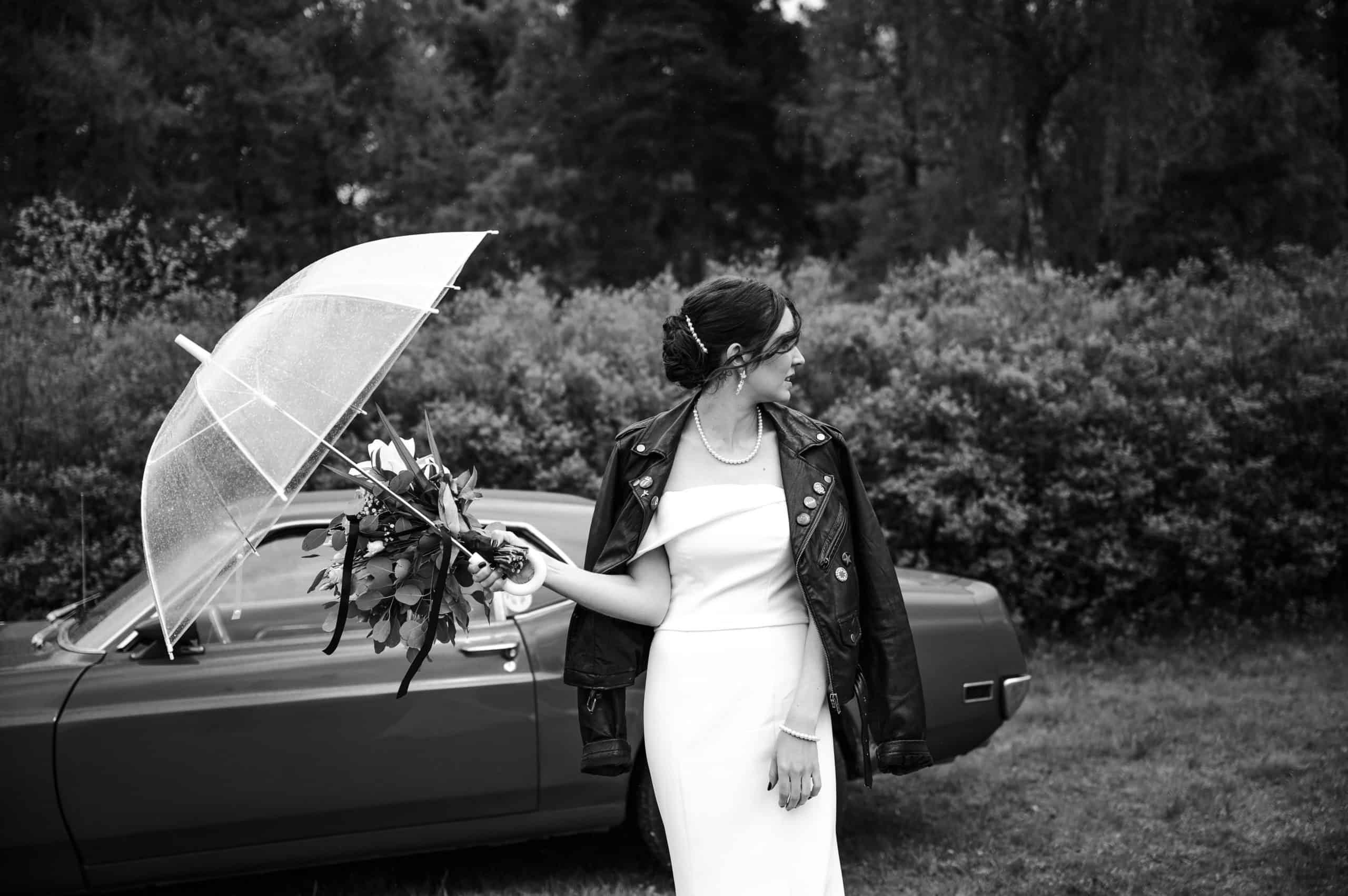 Kvinne med paraply og blomster ved bil i regnfullt maibryllup i Hønefoss.