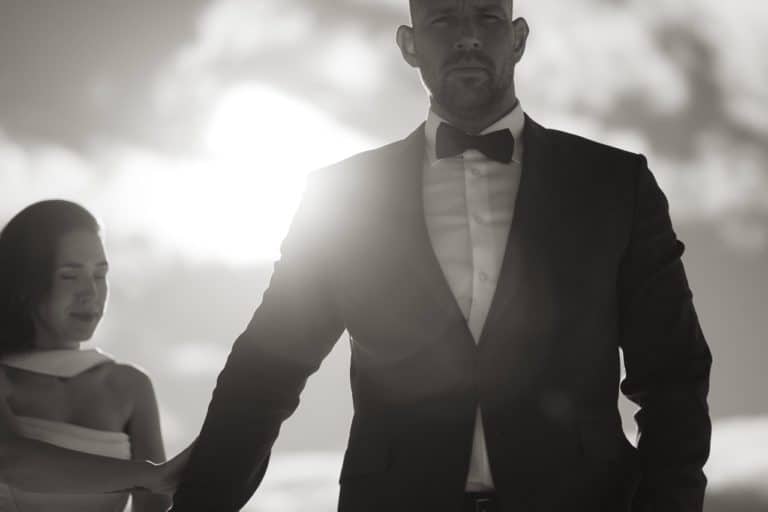 Elegant mann i svart dress og sløyfe med kvinne i bakgrunnen.