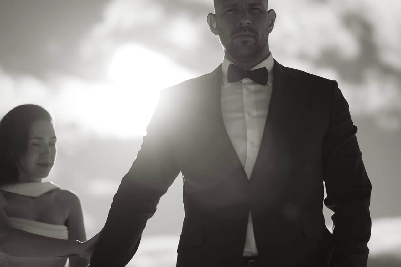 Elegant mann i svart dress og sløyfe med kvinne i bakgrunnen.