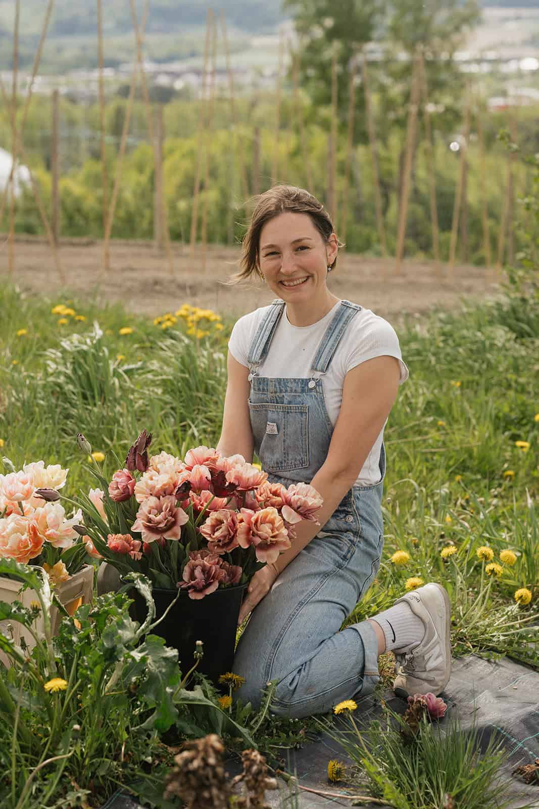 Kvinne plukker blomster i en blomsterhage med grønne trær og åker i bakgrunnen.