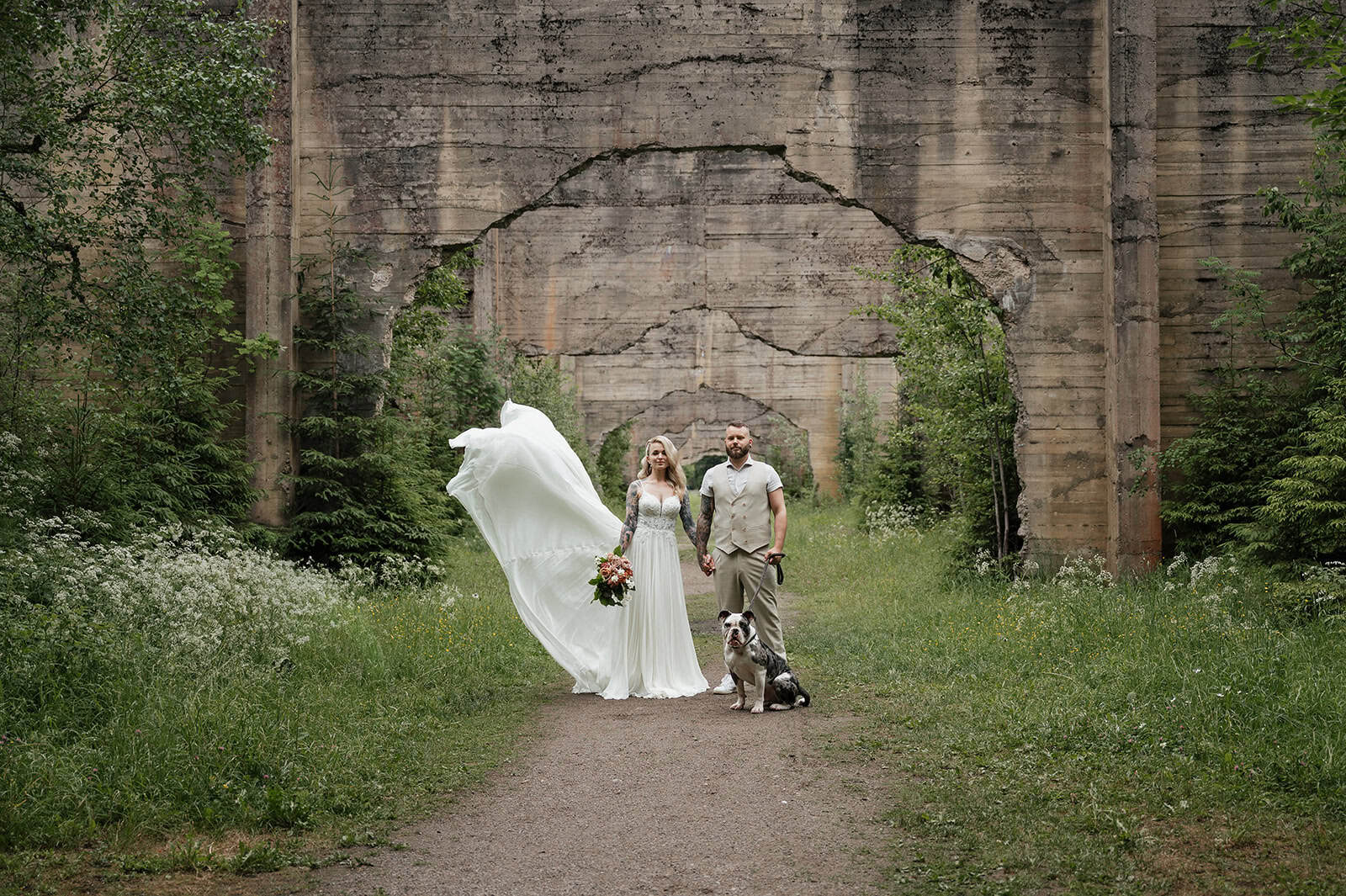 Bryllupsfoto av et par med hund foran en gammel bro i naturen.