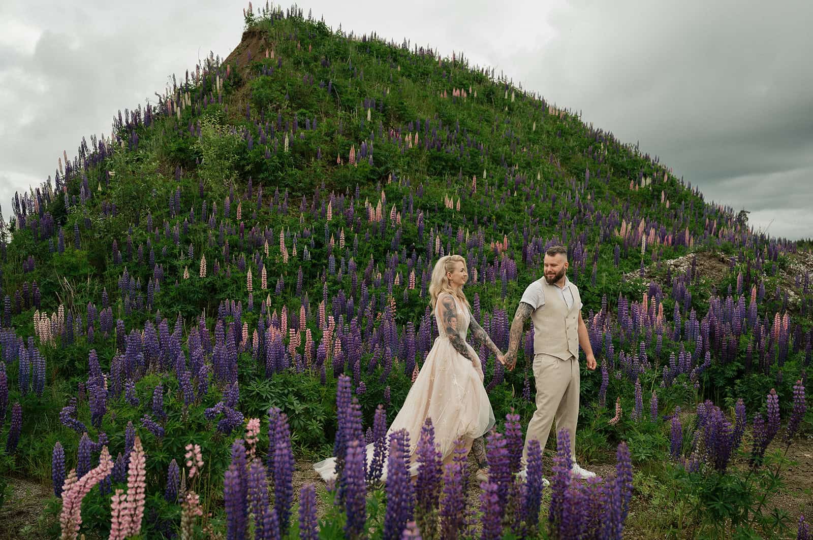 Par av mennesker i brudekjole og dress holder hender i blomsterfelt foran fjell.